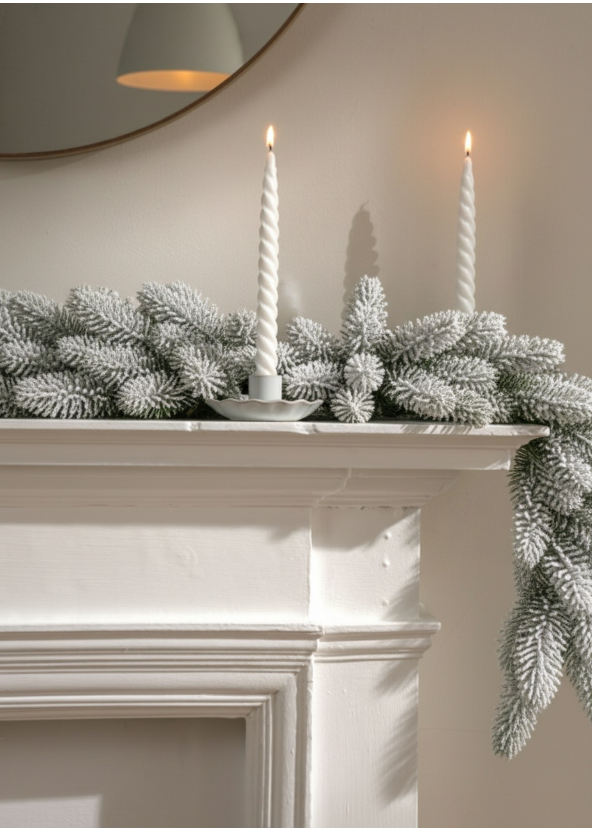 TRULY LIFESTYLE CHRISTMAS SNOWY GARLAND Decorative mantel with white candles and frosted greenery against a neutral wall.