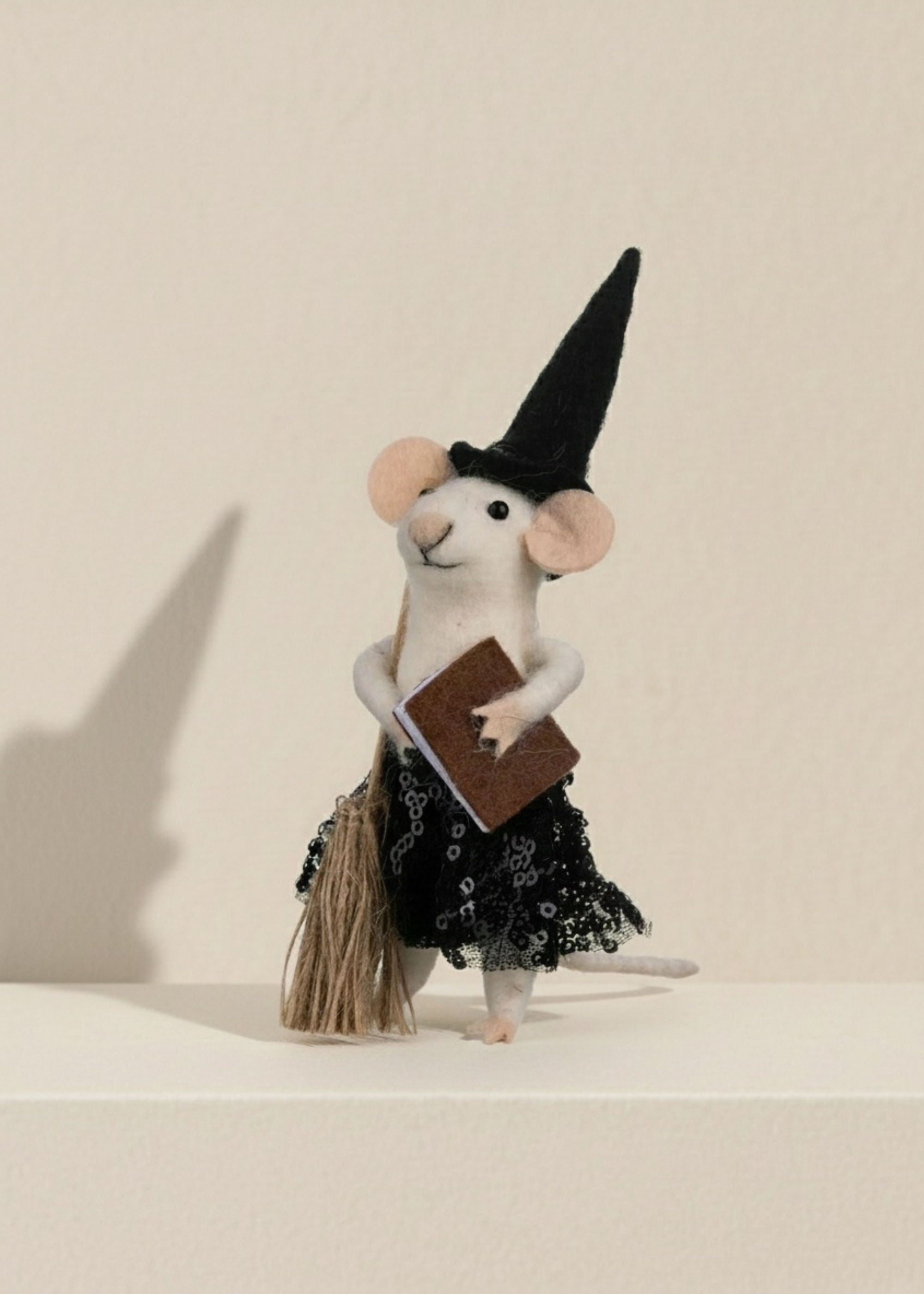 Stuffed mouse toy dressed as a witch holding a book and a broomstick on a beige background by Truly Lifestyle