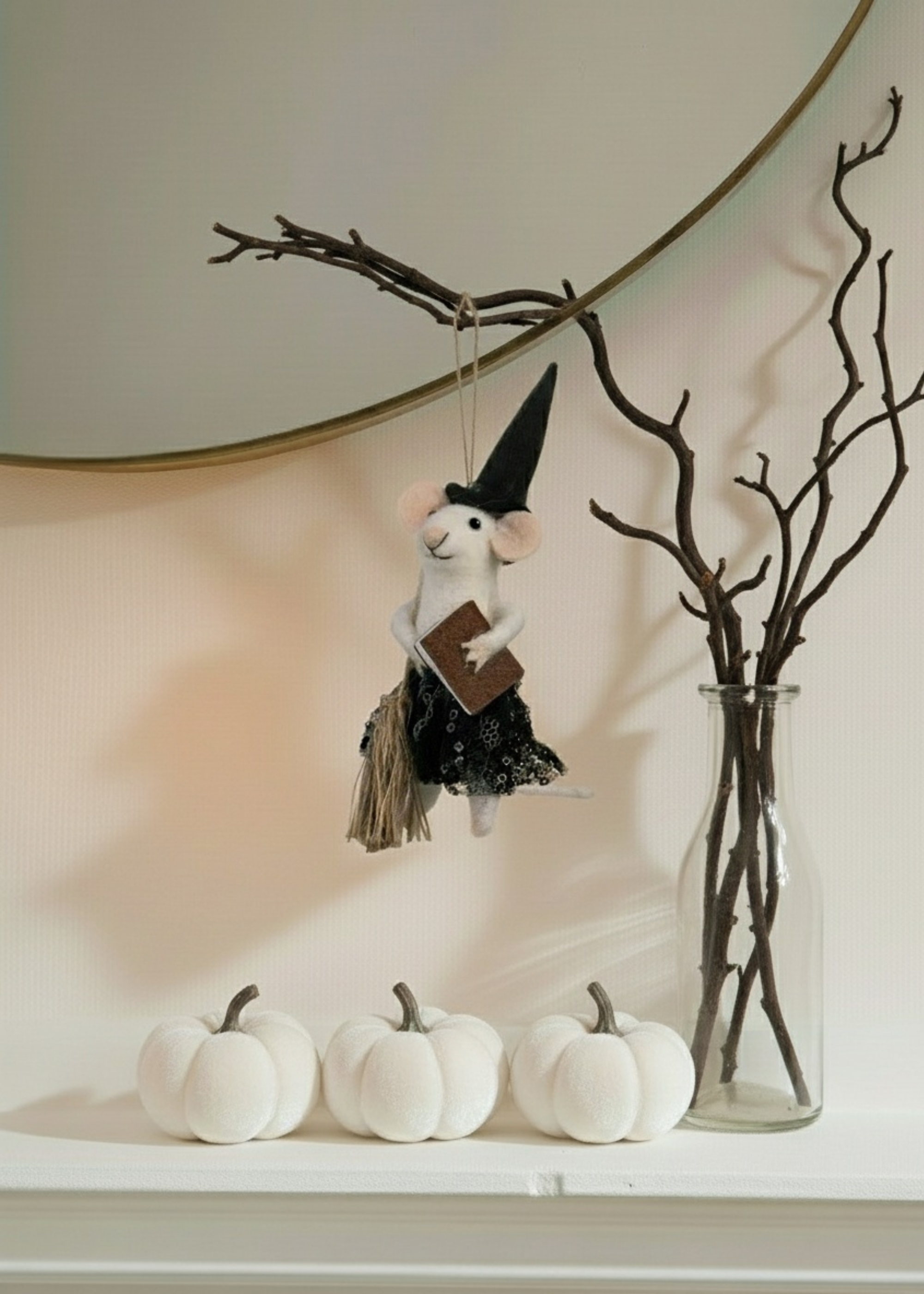Decorative mouse in a witch costume hanging from a branch with white pumpkins and a vase of branches on a neutral background.