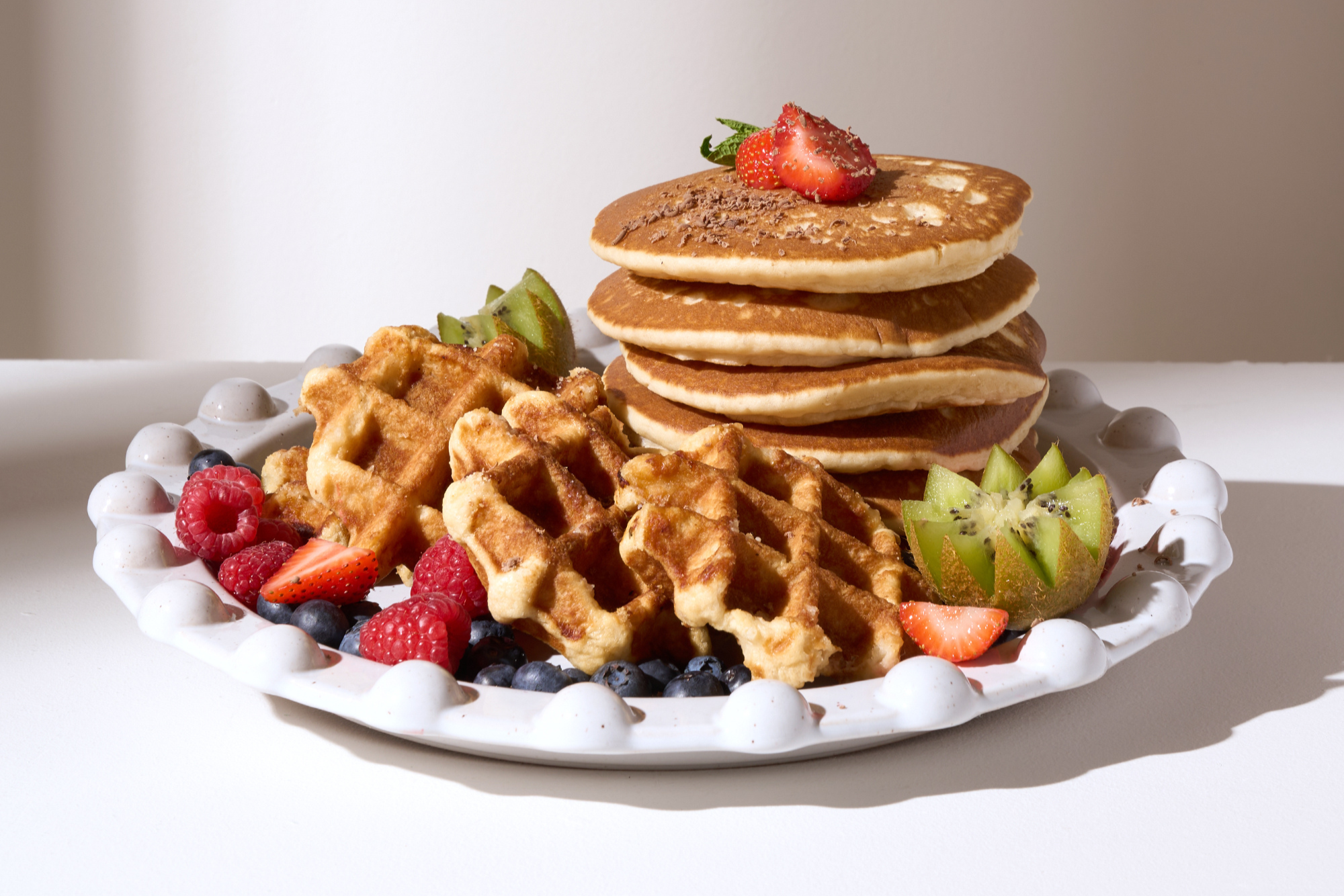 TRULY LIFESTYLE PALE GREY ROUND POM POM EDGED SERVING PLATTER FILLED WITH PANCAKES, WAFFLES AND FRUIT