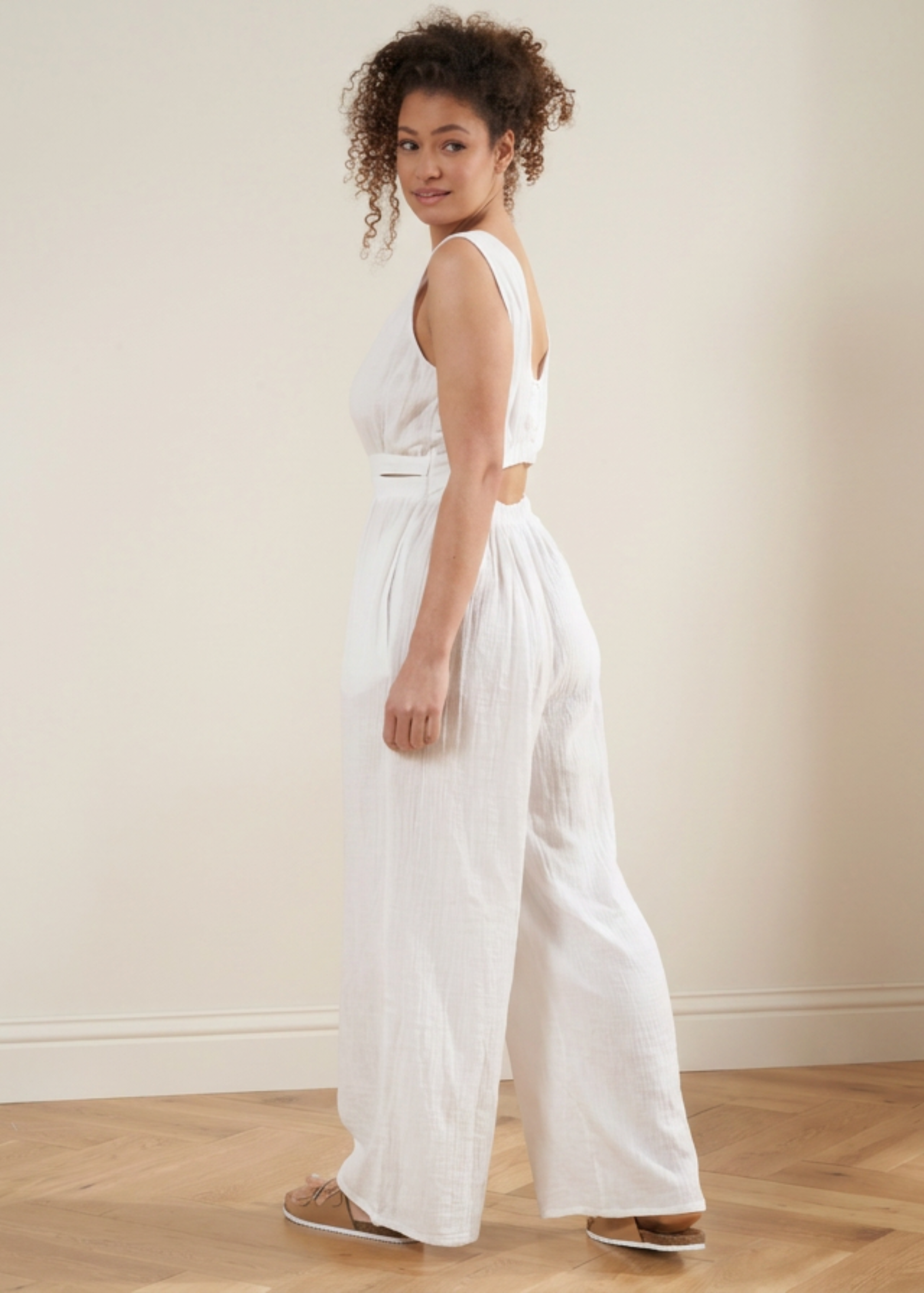 A woman wearing a white sleeveless wrap-style jumpsuit with a cutout at the waist and wide legs, standing in a minimalist room with a beige wall and light wood floor.