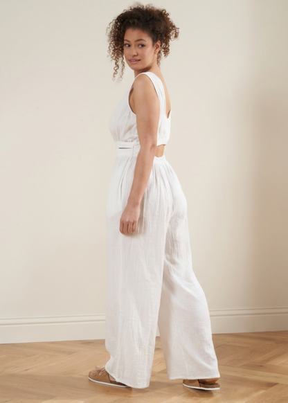 A woman wearing a white sleeveless wrap-style jumpsuit with a cutout at the waist and wide legs, standing in a minimalist room with a beige wall and light wood floor.